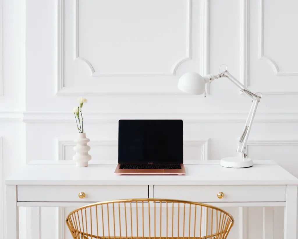 All white office space with a laptop, desk lamp and wicker chair.
